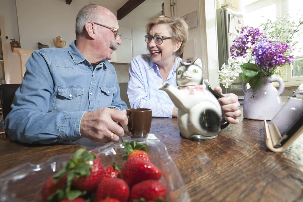 Een oudere man en vrouw zitten aan een houten tafel, lachend en houden mokken vast. Er staan een katvormige theepot en vazen met bloemen op tafel, samen met een bakje verse aardbeien op de voorgrond. Ze lijken te genieten van een luchtig moment, misschien samengebracht door een koppelaarster.