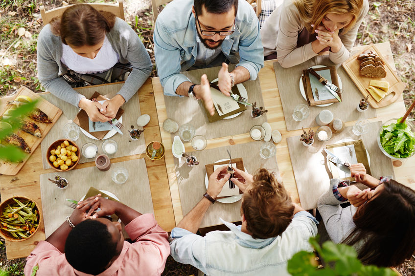 Bovenaanzicht van zes mensen die buiten rond een houten tafel zitten en een maaltijd delen. Ze lijken verwikkeld in een gesprek, met eten als brood, salade en aardappelen op tafel. De setting is ontspannen en gemeenschappelijk en bevordert een gevoel van verbondenheid, vergelijkbaar met wat het relatiebureau nastreeft bij het faciliteren van een duurzame relatie.