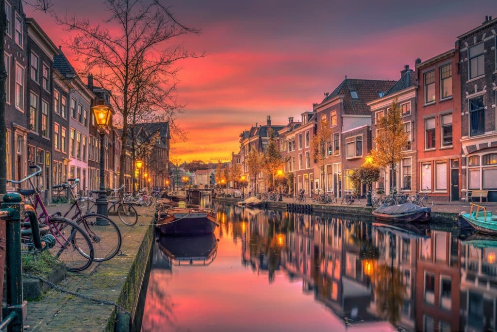 Een pittoresk kanaal in een Europese stad is omzoomd met traditionele bakstenen gebouwen. De ondergaande zon werpt een levendige mix van roze, oranje en paarse tinten door de lucht. Kleine boten liggen aangemeerd langs het kalme water en weerspiegelen de prachtige kleuren erboven, waardoor een sfeer ontstaat die perfect is voor een romantische matchmaking-wandeling.