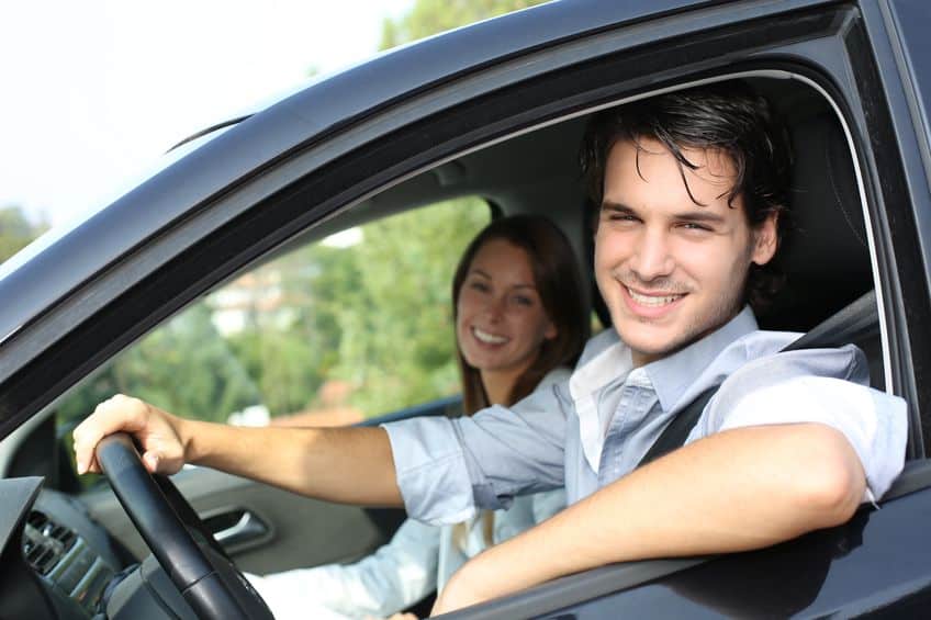 Een jonge man met kort donker haar zit op de bestuurdersstoel van een auto en lacht naar de camera. Op de passagiersstoel zit een vrouw met lang bruin haar, eveneens glimlachend. Het open autoraam onthult groen op de achtergrond en legt een moment vast dat de belichaming is van een succesvolle matchmaking die leidt tot een duurzame relatie.