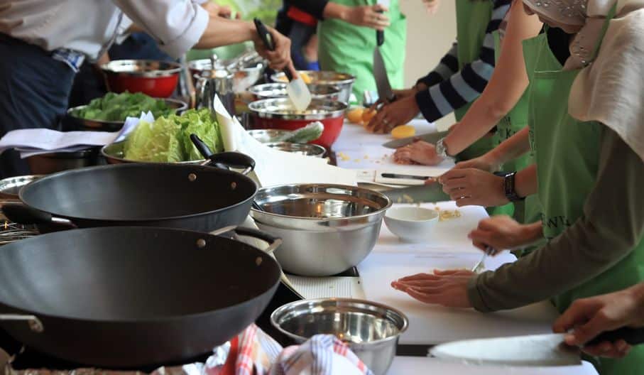 Een groep mensen met groene schorten verzamelt zich rond een aanrecht en is actief aan het koken. Diverse groenten, mengkommen en kookgerei staan verspreid. Sommige individuen hakken ingrediënten, terwijl anderen gerechten mixen en bereiden - het is een culinaire matchmaker die een duurzame relatie met voedsel creëert.