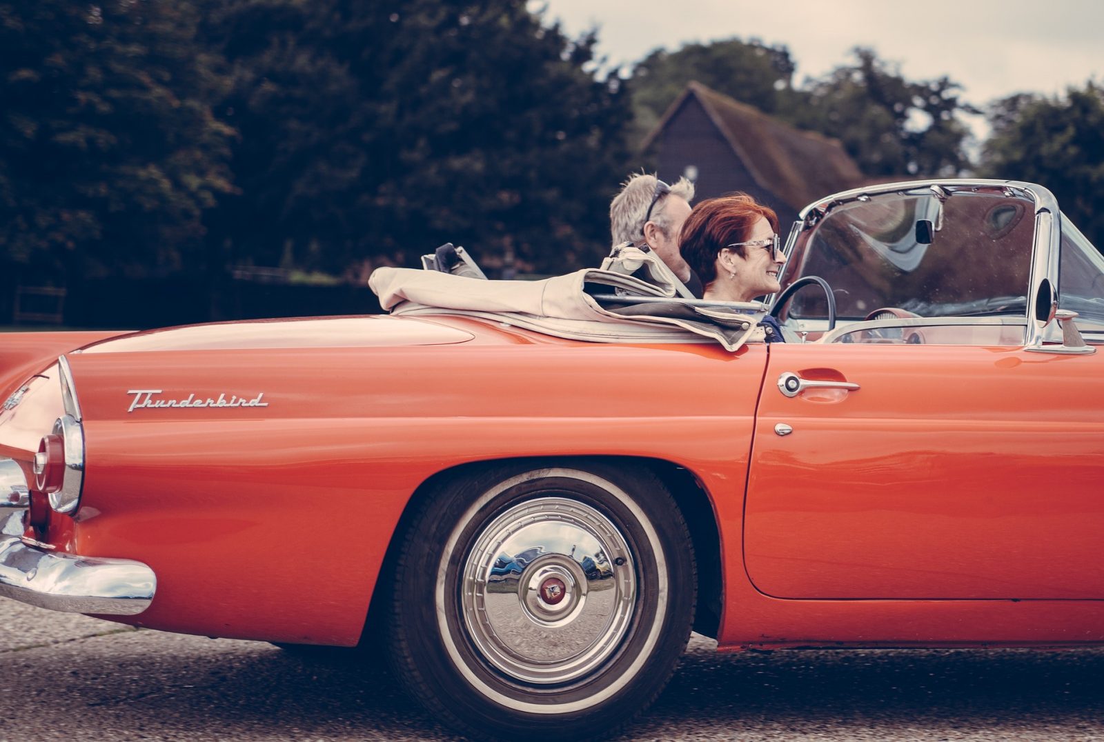 Een ouder echtpaar geniet van een ritje in een klassieke oranje Thunderbird-cabriolet met het dak naar beneden, rijdend door een schilderachtig landelijk gebied. De man rijdt terwijl de vrouw lacht, met bomen en een huis op de achtergrond zichtbaar – een beeld van wat kan bloeien uit verbindingen die tot stand zijn gekomen via een duurzame relatie.
