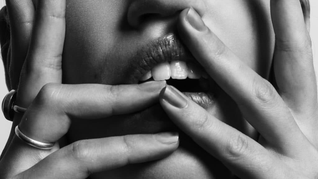 Zwart-wit close-up van het gezicht van een persoon met de handen die de lippen aanraken. Verschillende vingers, versierd met ringen, worden rond de mond geplaatst, waardoor nagels en tanden zichtbaar worden. Het intieme shot brengt een diepte van emotie over die lijkt op de nuances die een ervaren koppelaarster begrijpt.