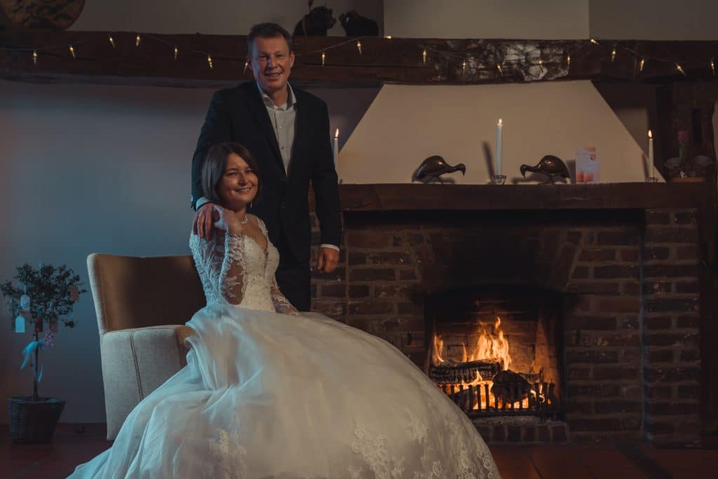 Een stel poseert in een elegante setting voor een relatiebemiddelingsadvertentie. De vrouw, gekleed in een witte trouwjurk, zit op een stoel met haar hand op de arm. De man, gekleed in pak, staat achter haar met zijn hand op haar schouder. Ze staan voor een verlichte bakstenen open haard.