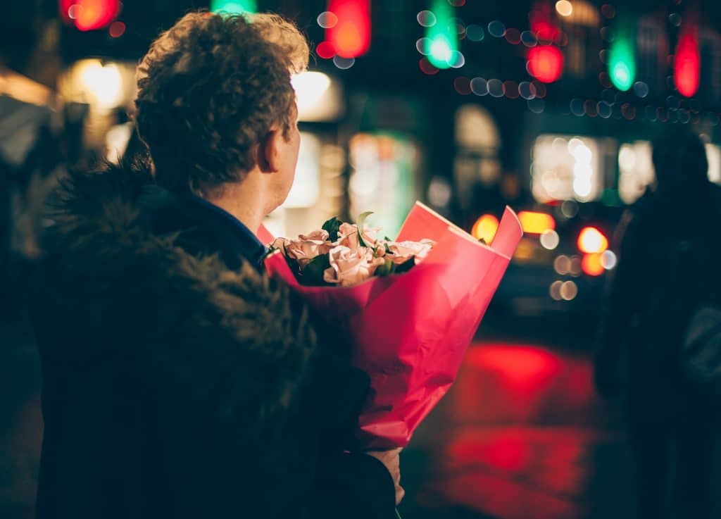 Een persoon met krullend haar en een met bont afgezette jas draagt 's nachts in een stadsstraat met een boeket roze bloemen in rood papier gewikkeld. De straat is versierd met kleurrijke lichtjes, wat suggereert dat ze via een matchmaking-service een speciaal iemand kunnen ontmoeten. Wazige figuren en voertuigen zijn op de achtergrond.