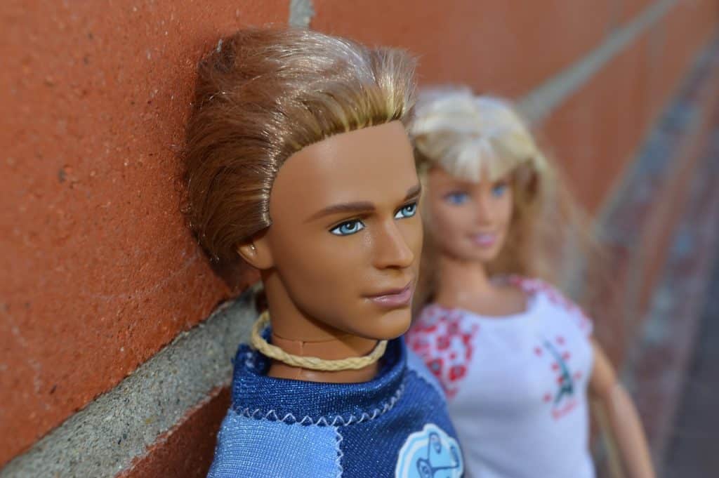 Close-up van twee modepoppen die tegen een bakstenen muur leunen. De mannelijke pop, met een gebruinde huid en blond haar, staat op de voorgrond en draagt een blauw topje. De vrouwelijke pop, met blond haar en een lichte huid, staat op de achtergrond en roept in haar witte overhemd met bloemenmotief een aura van duurzame relatie op.