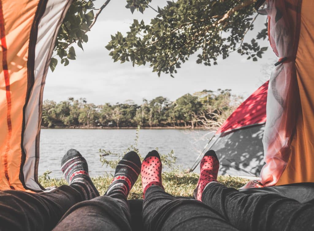 Uitzicht vanuit een tent met twee paar voeten in kleurrijke sokken die uitsteken. De tent staat vlakbij een watermassa omringd door bomen en groen, als een serene schuilplaats, perfect voor het bevorderen van een duurzame relatie. Op de achtergrond is onder een heldere, gedeeltelijk bewolkte hemel een andere tent zichtbaar.