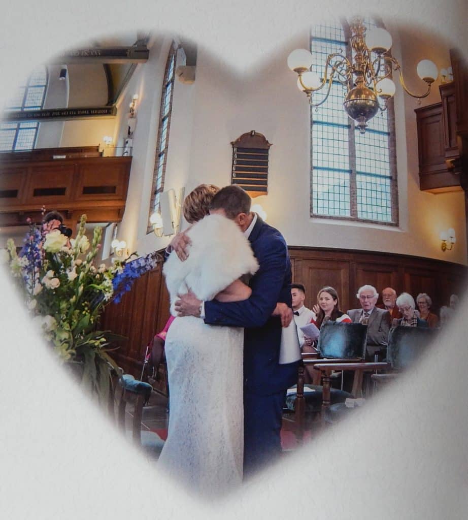 Een bruid en bruidegom, bijeengebracht door een matchmaking-datingbureau, delen een omhelzing in een hartvormige omlijsting in een kerk. De bruid draagt een witte jurk en een bontstola, terwijl de bruidegom een blauw pak draagt. Gasten zitten op de achtergrond, samen met bloemen en houten lambrisering aan de muren.