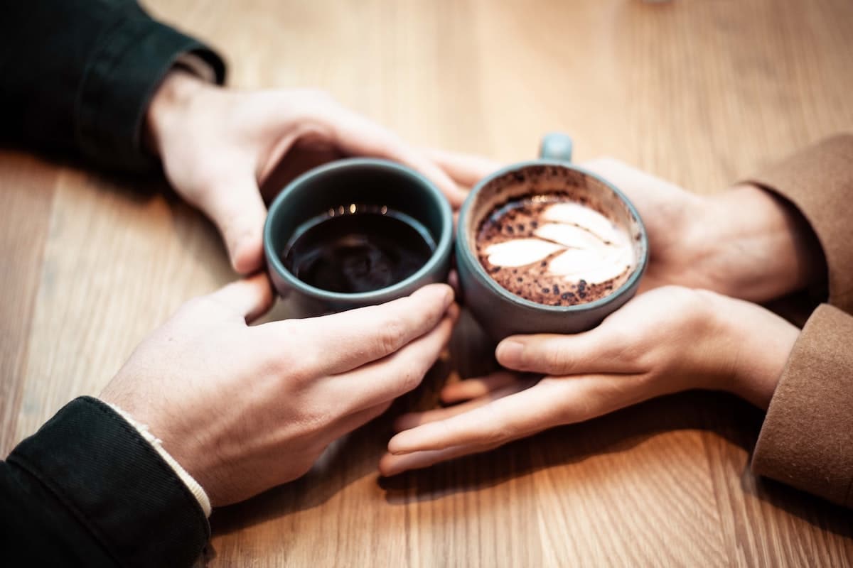 Twee mensen rammelen keramische mokken tegen elkaar boven een houten tafel. In de ene mok zit zwarte koffie, in de andere een schuimige cappuccino met daarop latte art. De handen zijn dichtbij elkaar, wat een moment van warmte en verbondenheid benadrukt, wat doet denken aan de vonken die vaak te zien zijn bij succesvolle matchmaking-ontmoetingen.