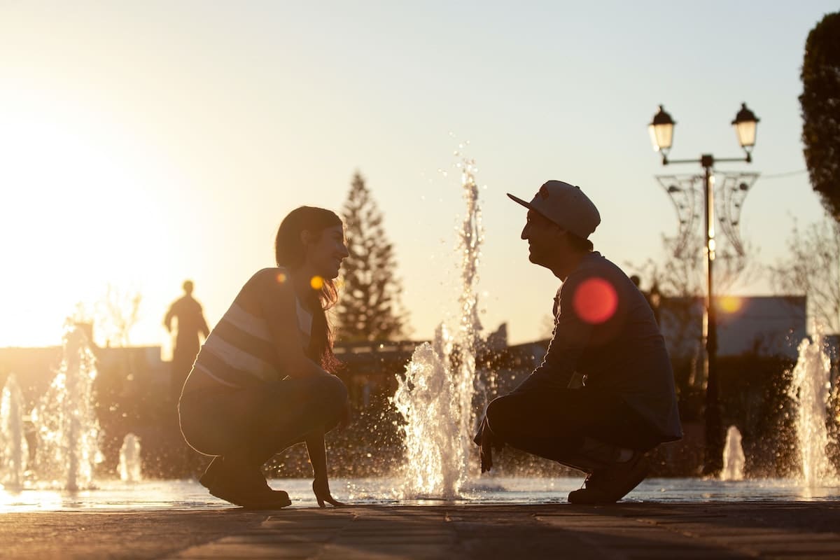 Een man en een vrouw hurken en kijken elkaar aan voor een reeks waterfonteinen bij zonsondergang, misschien bespreken ze hun reis naar het vinden van een duurzame relatie. De zon staat laag aan de hemel, waardoor een warme gloed ontstaat en silhouetten van het stel ontstaan. Op de achtergrond loopt een persoon voorbij, waarbij ook bomen en lantaarnpalen zichtbaar zijn.