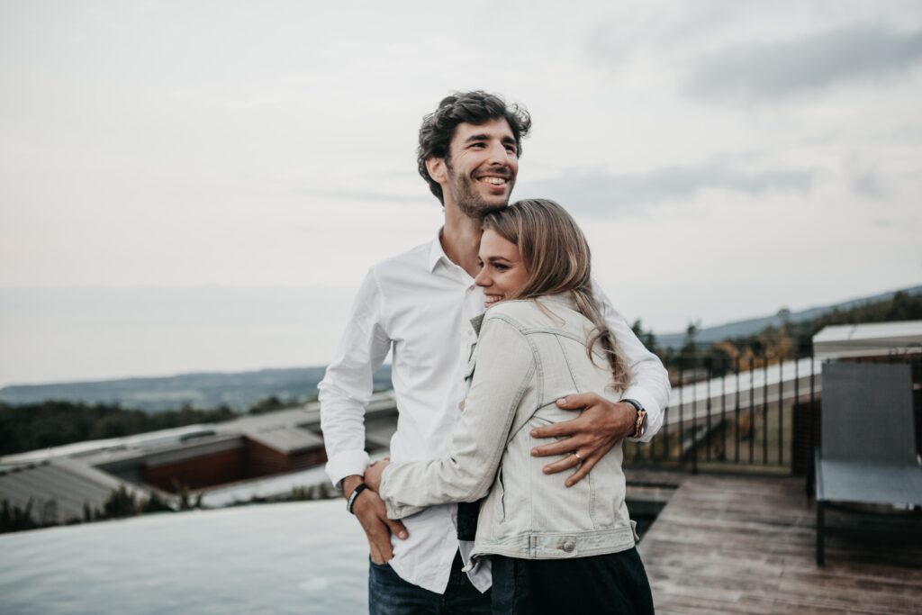 Een man en een vrouw staan elkaar op een dakterras te omhelzen. De vrouw, gekleed in een licht spijkerjasje, glimlacht terwijl ze zich tegen de man aan leunt. De man, in een wit overhemd, kijkt weg en glimlacht. Ze delen een moment dat wordt gevangen door het schilderachtige uitzicht op gebouwen en bomen – een bewijs van hun reis door middel van relatiebemiddeling om een duurzame relatie op te bouwen.
