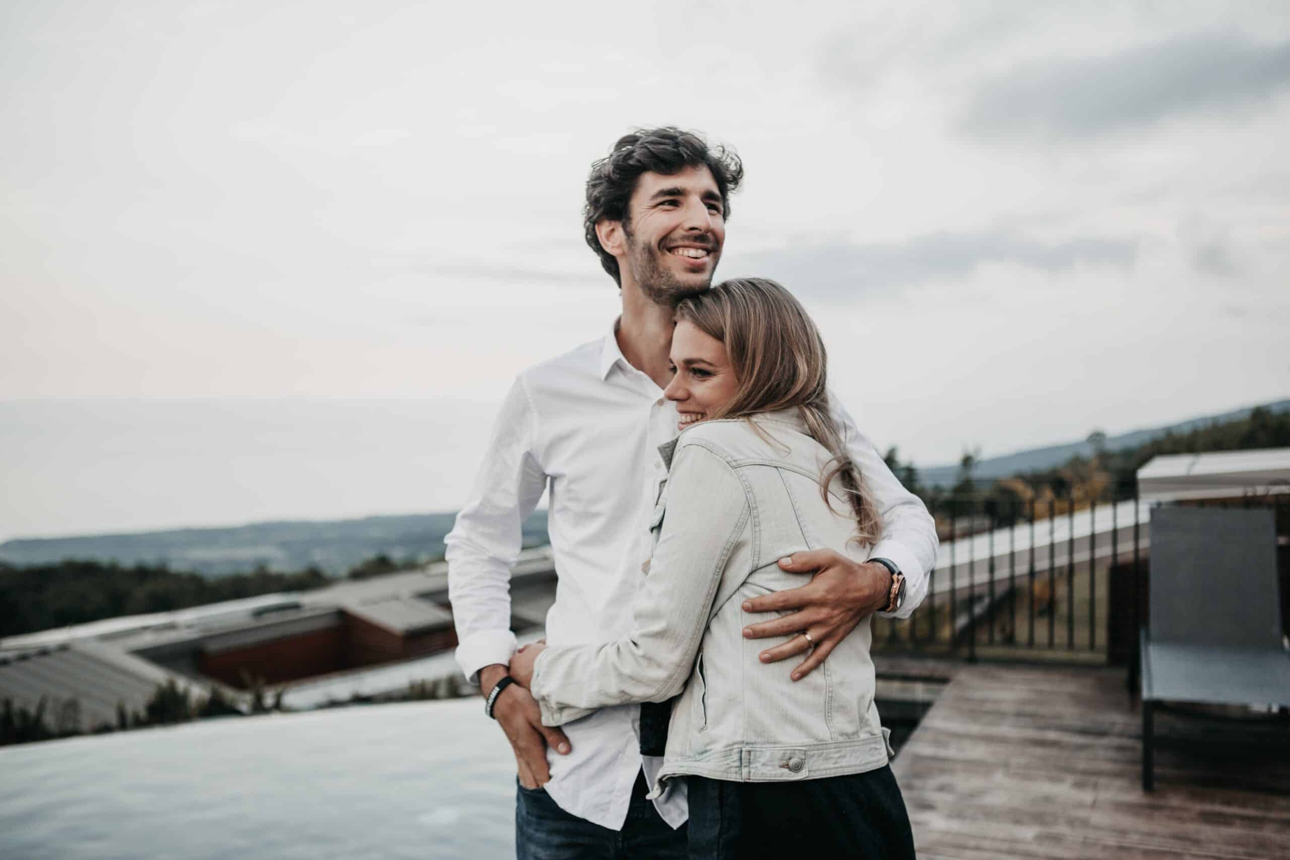 Een man en een vrouw staan elkaar op een dakterras te omhelzen. De vrouw, gekleed in een licht spijkerjasje, glimlacht terwijl ze zich tegen de man aan leunt. De man, in een wit overhemd, kijkt weg en glimlacht. Ze delen een moment dat wordt gevangen door het schilderachtige uitzicht op gebouwen en bomen – een bewijs van hun reis door middel van relatiebemiddeling om een duurzame relatie op te bouwen.