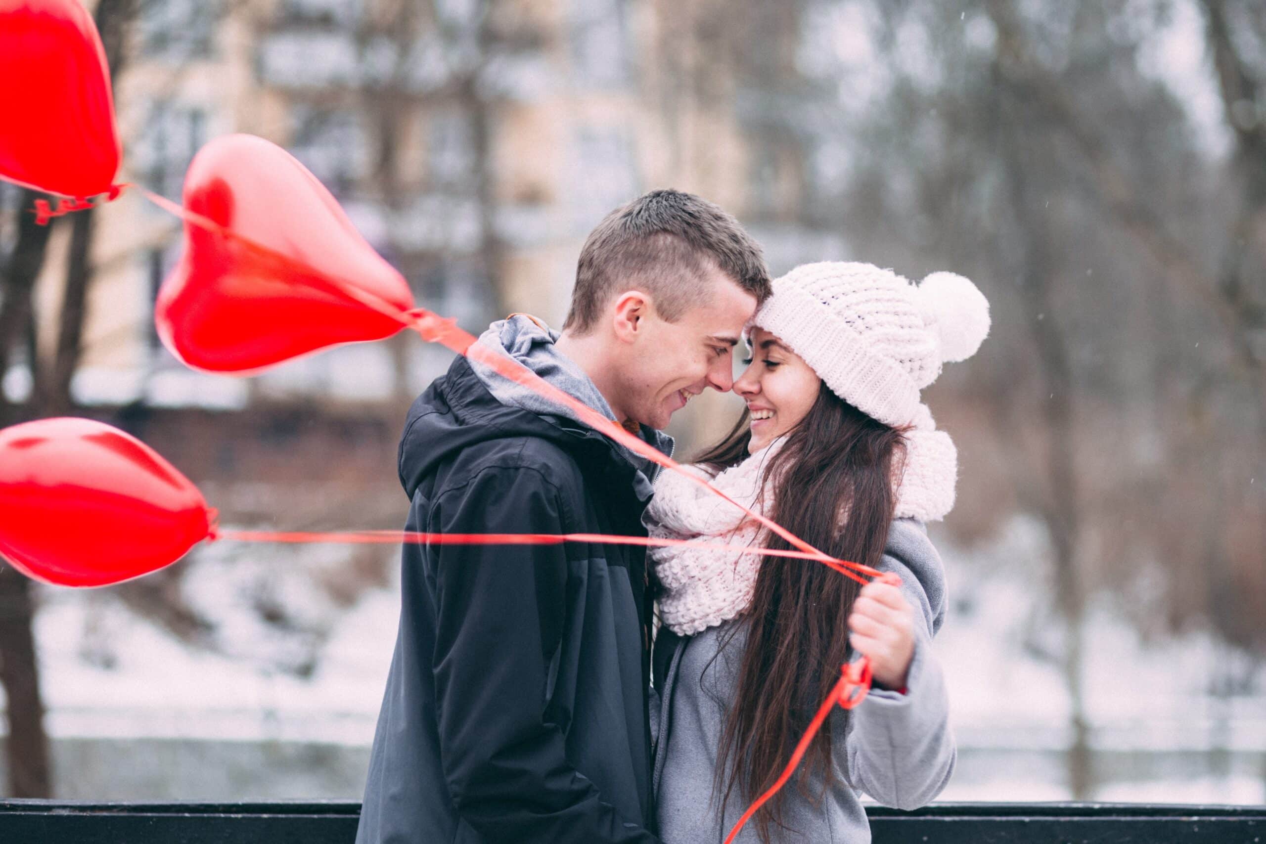 Een jong stel staat buiten dicht bij elkaar, glimlachend en elkaars voorhoofd aanrakend. De vrouw, die een witte gebreide muts en sjaal draagt, houdt verschillende rode hartvormige ballonnen vast. Dit tedere moment weerspiegelt de vreugde die wordt gecreëerd door relatiebemiddeling tegen een besneeuwde achtergrond van vage bomen en gebouwen.