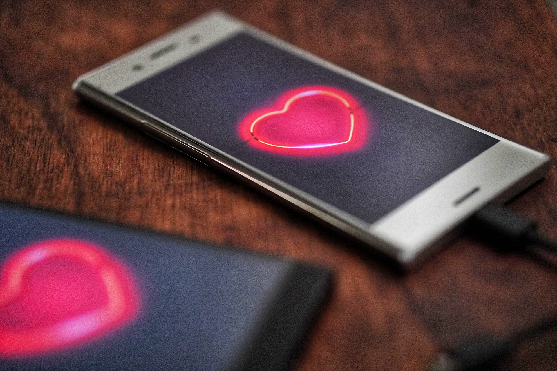 Een smartphone met een gloeiend neonhart op het scherm rust op een houten oppervlak, wat de belofte van een duurzame relatie symboliseert. Een andere smartphone met een wazig scherm is gedeeltelijk zichtbaar op de voorgrond. Beide telefoons zijn verbonden via oplaadkabels.