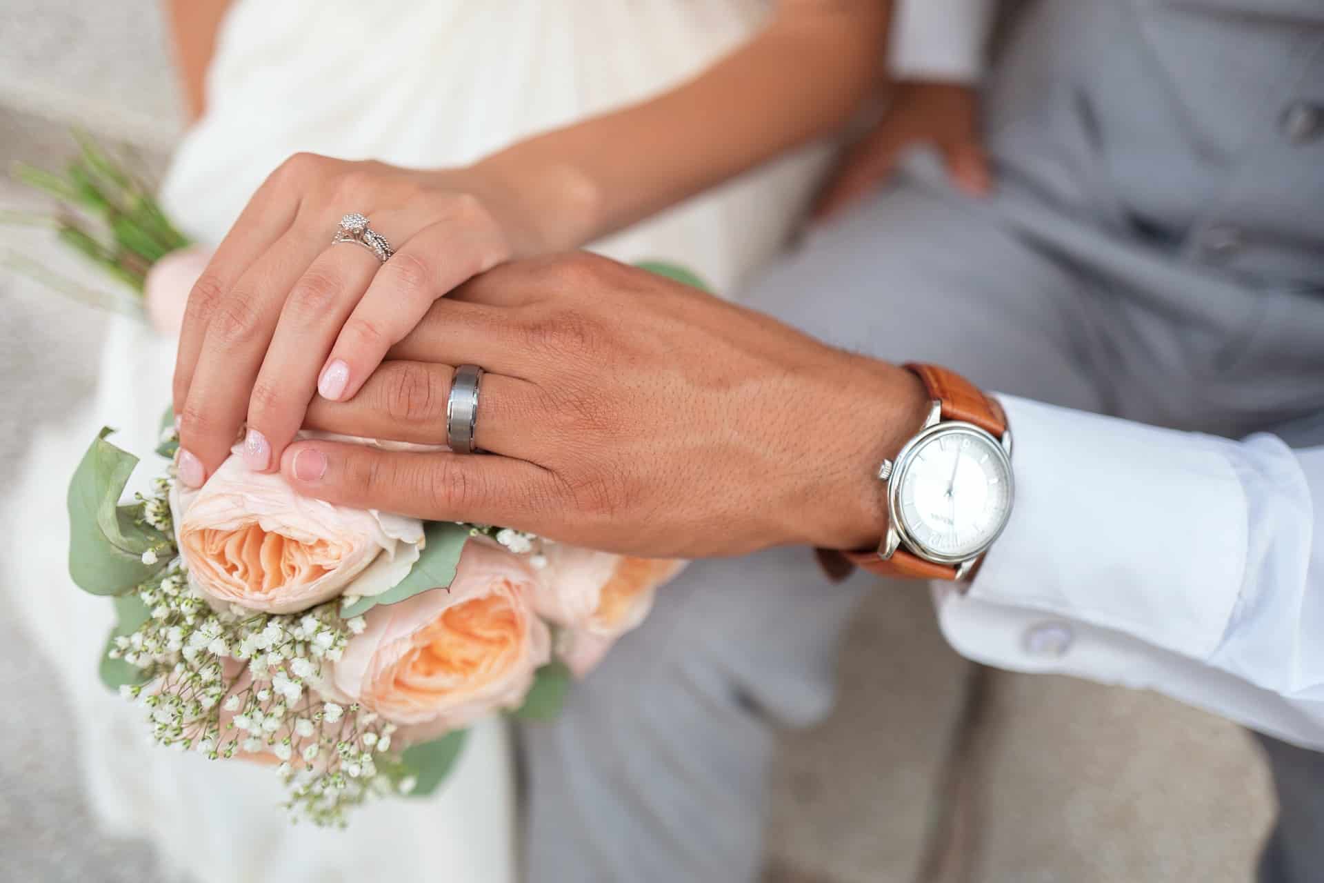 Close-up van de handen van een stel op een boeket perzikrozen en gipskruid. De hand van de vrouw, met een diamanten verlovingsring, rust er bovenop. De hand van de man, die een zilveren trouwring en polshorloge draagt, houdt de hare zachtjes vast. Beiden dragen lichtgekleurde kleding, met dank aan hun toegewijde matchmaking-service.