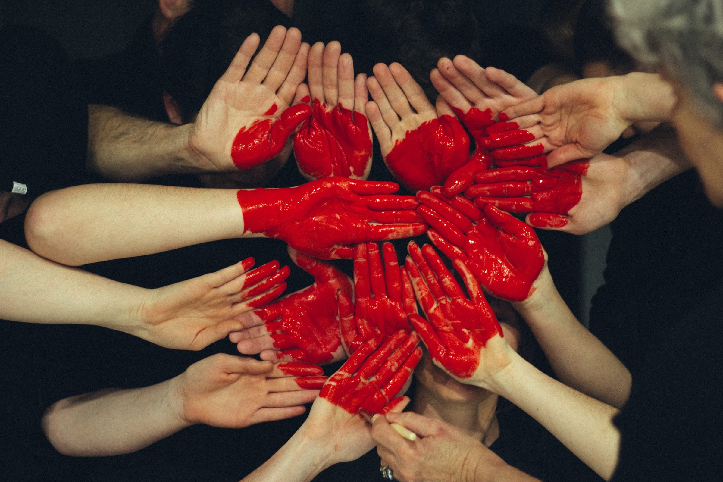 Een groep handen, waarvan sommige rood geverfd zijn, zijn in een cirkel gerangschikt met roodgeverfde handen die in het midden een hartvorm vormen. Het beeld symboliseert eenheid en samenwerking, tegen een donkere achtergrond – een suggestieve weergave van hoe een datingbureau duurzame relaties bevordert.