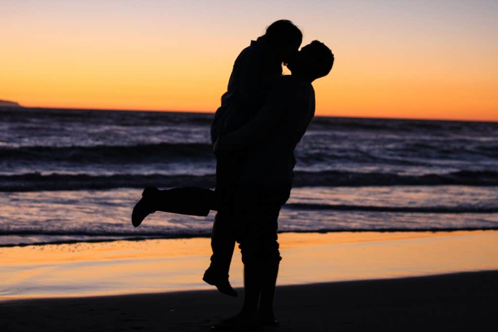 Een silhouet van twee mensen die elkaar omhelzen op een strand bij zonsondergang, opgeheven in een hartstochtelijke kus. De lucht is beschilderd met tinten oranje en paars, terwijl golven zachtjes op de achtergrond neerstorten – een perfect moment samengesteld door een matchmaking-service die zich inzet voor het bevorderen van duurzame relaties.