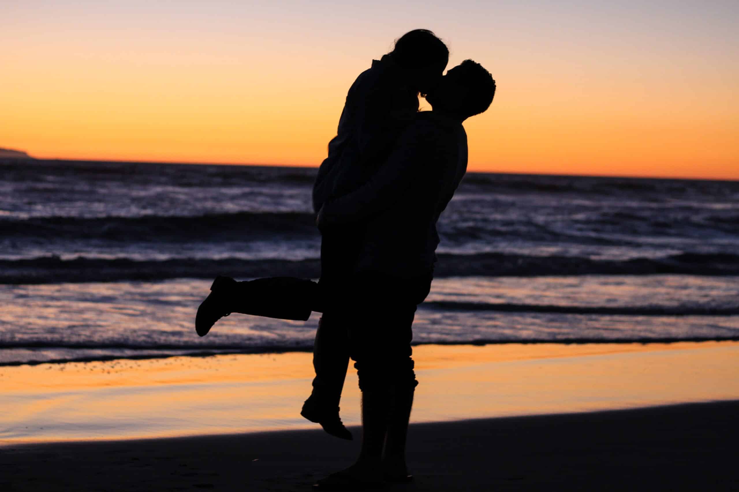 Een silhouet van twee mensen die elkaar omhelzen op een strand bij zonsondergang, opgeheven in een hartstochtelijke kus. De lucht is beschilderd met tinten oranje en paars, terwijl golven zachtjes op de achtergrond neerstorten – een perfect moment samengesteld door een matchmaking-service die zich inzet voor het bevorderen van duurzame relaties.