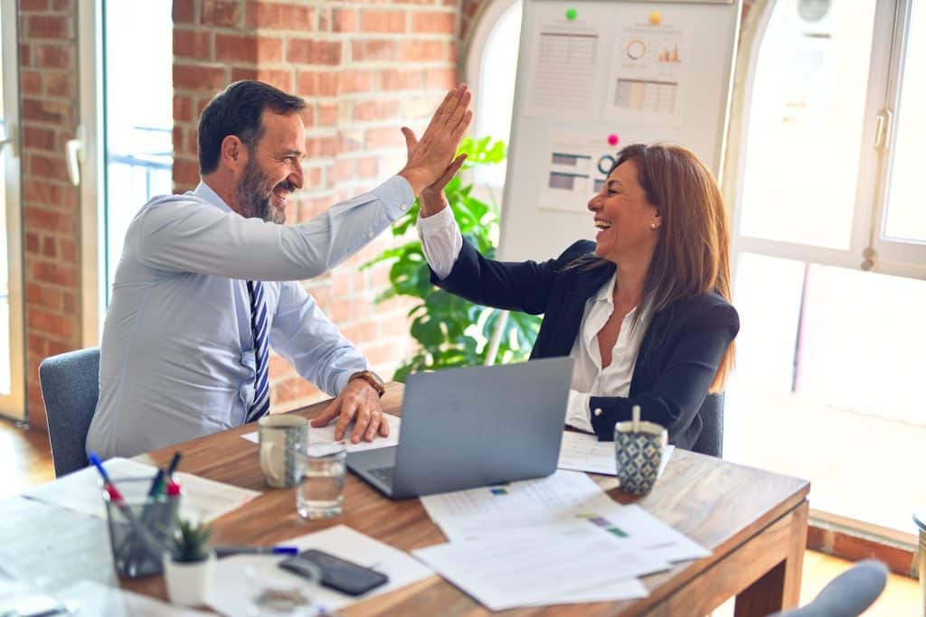 Een man en een vrouw in zakelijke kleding delen een high-five in een kantooromgeving, wellicht ter ere van hun succesvolle werk bij een relatiebureau. Ze zitten aan een houten bureau met een laptop, documenten, een koffiemok en een herbruikbare beker. Op de achtergrond zijn een bakstenen muur en een whiteboard met grafieken zichtbaar.