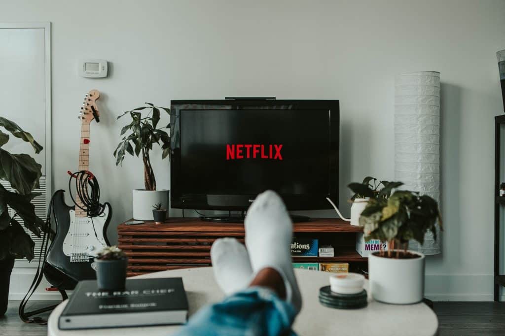 Een persoon ligt met de voeten omhoog op een bank en kijkt naar een tv waarop het Netflix-logo staat. De kamer is voorzien van potplanten, een gitaar, een boekenplank en een moderne inrichting. De algehele sfeer is gezellig en ontspannen, perfect om te settelen nadat je je hebt aangemeld bij een matchmaking-service om die ene speciale persoon te vinden.