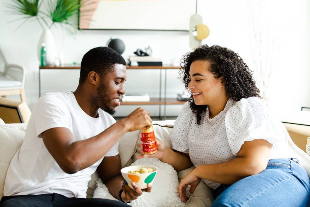 Een man en een vrouw zitten op een gezellige bank in een lichte, moderne woonkamer. De man houdt een kom eten vast en schenkt saus uit een fles, terwijl de vrouw, gekleed in een blouse met witte stippen, glimlacht en toekijkt. Het is duidelijk dat hun verbinding vakkundig tot stand is gebracht door een uitzonderlijke matchmaking-service. Op de achtergrond staan planten en decoratieve voorwerpen.