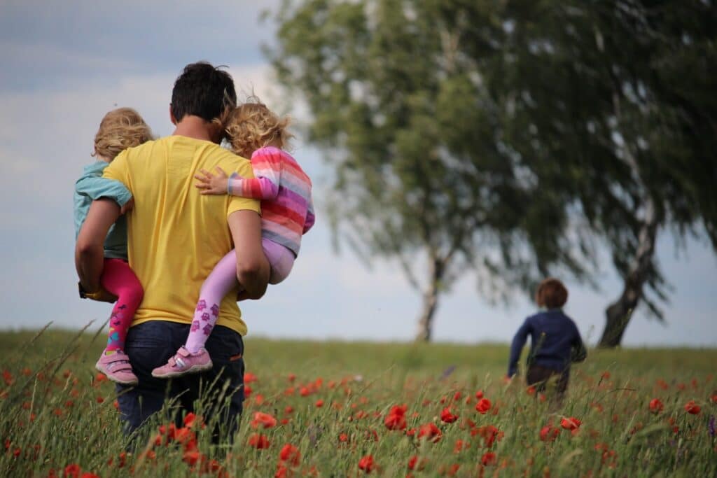 In een veld met rode klaprozen staat een persoon, gekleed in een geel shirt, met twee kinderen in zijn armen. Het ene kind draagt een blauw jasje en het andere kind is gekleed in een roze gestreepte outfit. Een ander kind, wellicht het resultaat van succesvolle matchmaking van een duurzame relatiedienst als het relatiebureau, loopt voorop. Op de achtergrond zijn bomen zichtbaar.