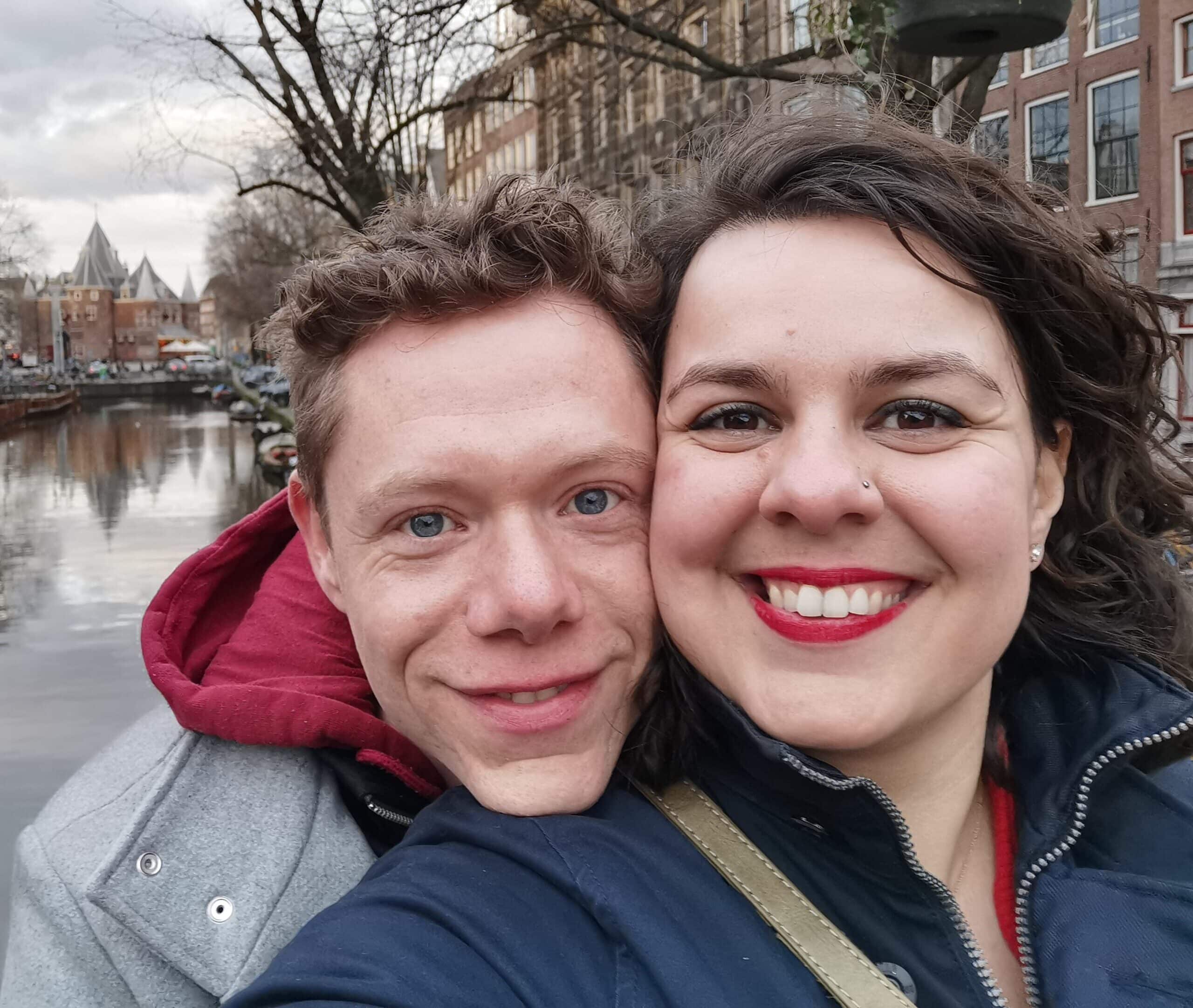 Succesverhalen - Start 2 Een glimlachend koppel maakt een close-up selfie langs een gracht in een stad met historische gebouwen op de achtergrond. De man heeft licht haar en draagt een grijze jas met een rode capuchon, terwijl de vrouw donker haar, rode lippenstift heeft en marineblauw gekleed is. Hun vrolijke uitdrukking doet vermoeden dat ze elkaar via een datingbureau hebben ontmoet.