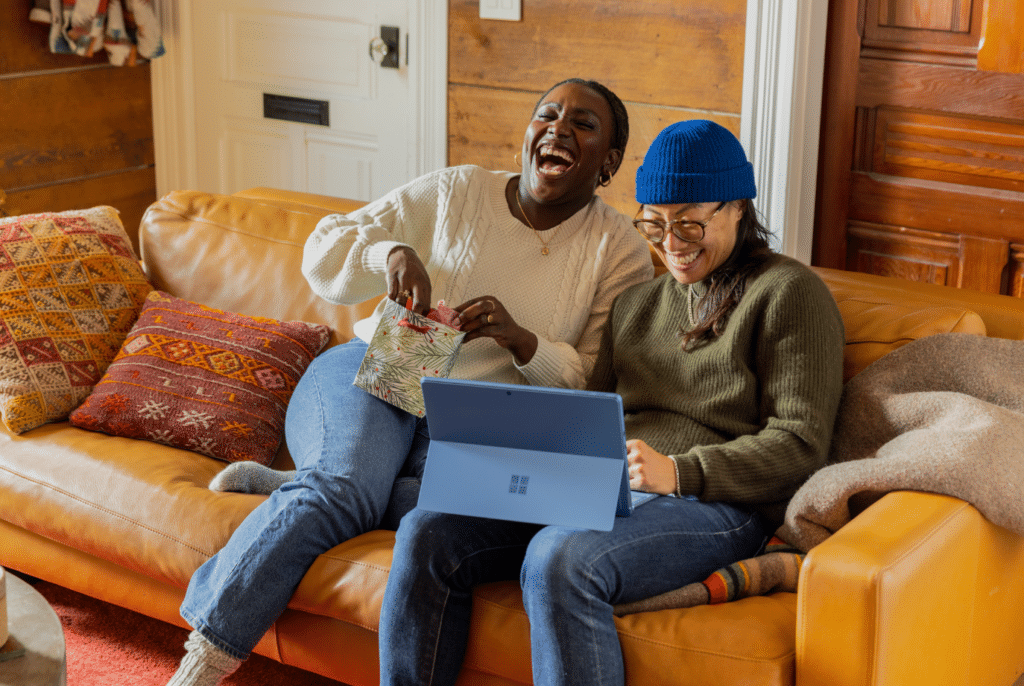 Twee mensen zitten op een lichtbruine leren bank in een gezellige kamer, lachend en glimlachend. De één maakt een cadeauzakje open, terwijl de ander, met een blauwe muts op, naar een laptop kijkt. Decoratieve kussens sieren de bank tegen houten wanden. Ze hebben elkaar ontmoet via een datingbureau en delen nu dit mooie moment in hun duurzame relatie.