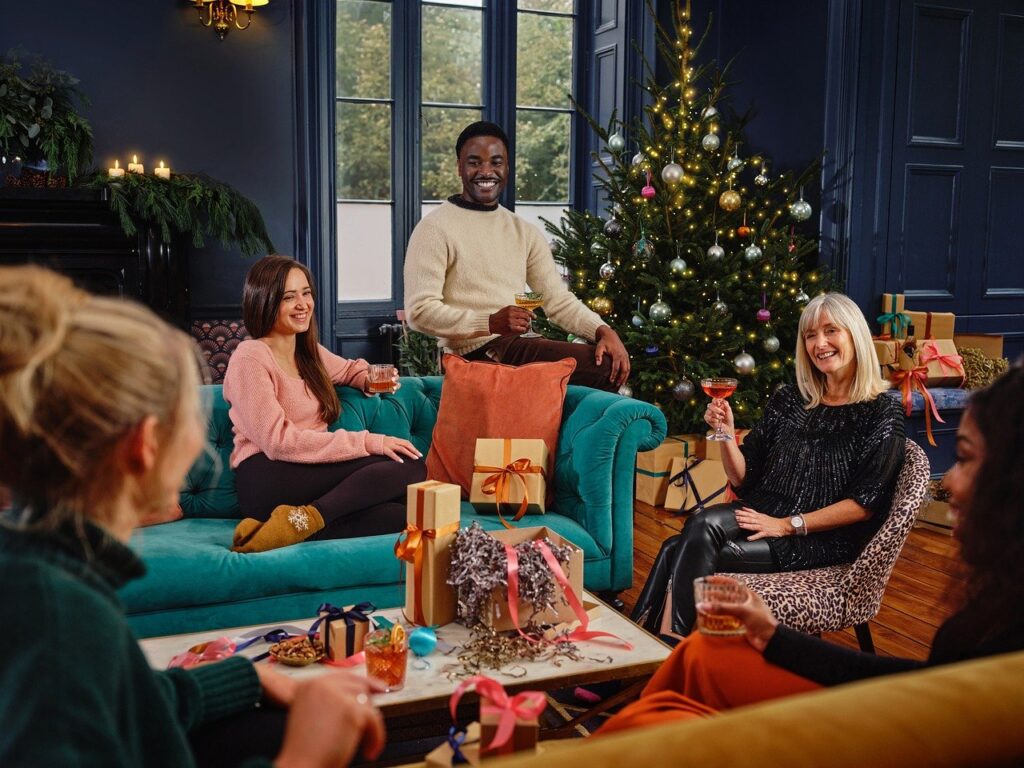 Een groepje van vijf mensen zit en staat rond een woonkamer, die voor Kerstmis versierd is met een boom en cadeautjes. Ze glimlachen en houden drankjes vast en lijken te genieten van een vakantiebijeenkomst georganiseerd door een eersteklas datingbureau. De kamer is gezellig, met blauwe muren en een feestelijke sfeer.