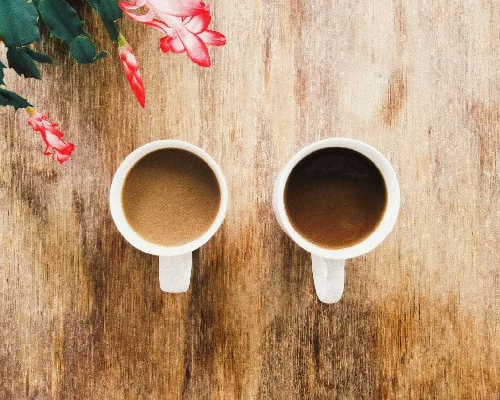Twee witte koffiemokken gevuld met koffie staan naast elkaar op een rustiek houten oppervlak. In de linkerbovenhoek van de afbeelding staan roze bloemen met groene bladeren, waardoor een warme en gezellige sfeer ontstaat, perfect voor het bespreken van zaken die verband houden met een duurzame relatie.