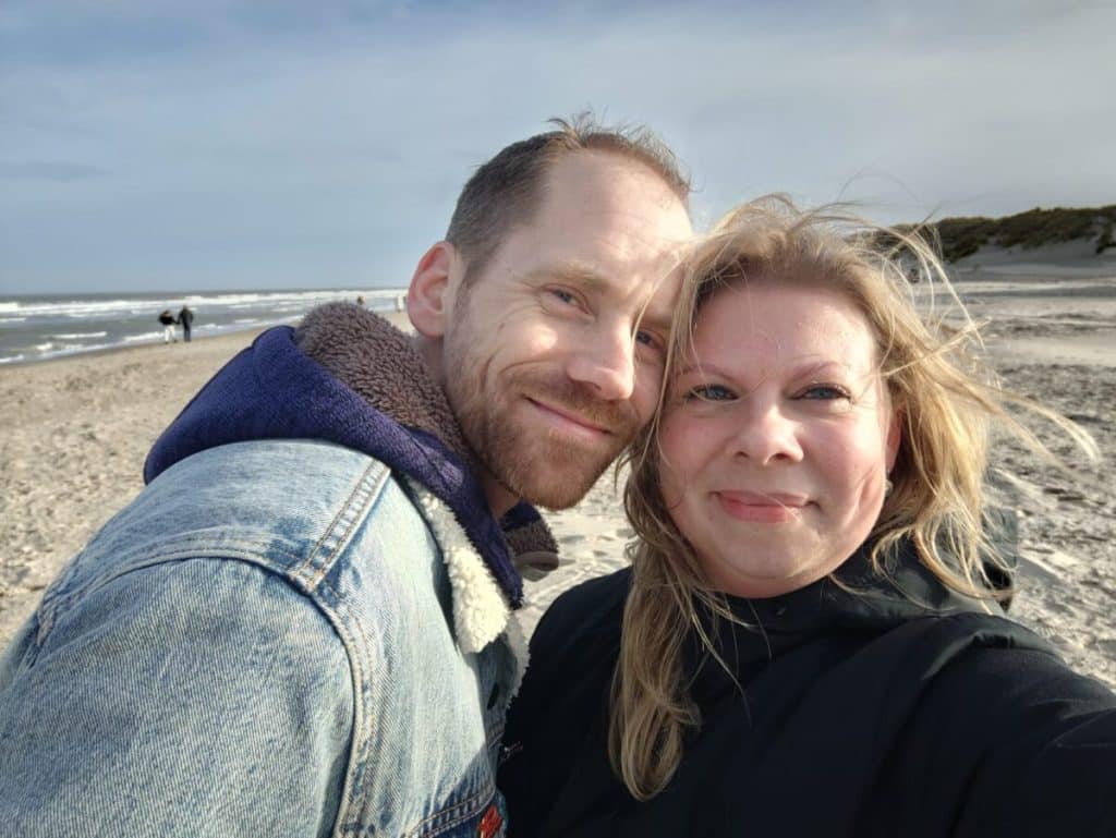 Een man en een vrouw glimlachen terwijl ze een selfie maken op het strand. De oceaan en de zandkust zijn zichtbaar op de achtergrond, met een bewolkte lucht erboven. Warm gekleed in jasjes suggereren hun vrolijke uitdrukkingen dat ze elkaar misschien via een datingbureau hebben ontmoet, terwijl het haar van de vrouw speels door de wind waait.