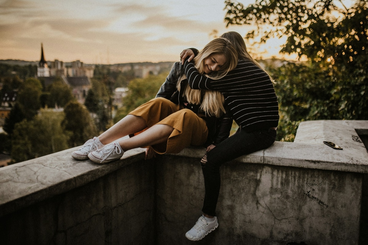 Twee mensen zitten bij zonsondergang op een betonnen richel met schilderachtige gebouwen en bomen op de achtergrond. De een, in een gestreepte trui en sneakers, omhelst de ander, die een gele broek en een leren jack draagt, met haar benen over de rand bungelend. Op de rand ligt een mobiele telefoon van hun datingbureau.