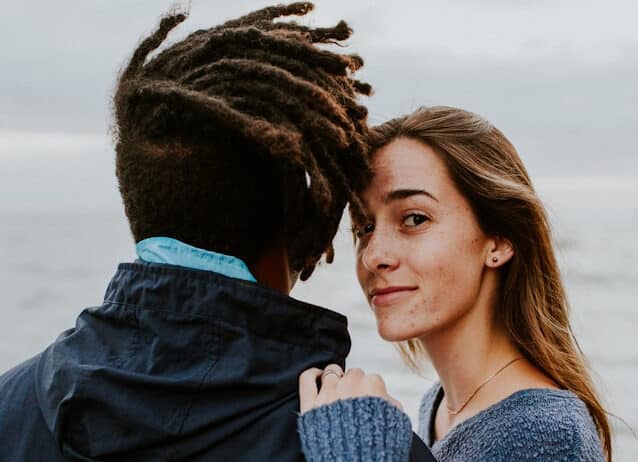 Een persoon met lang haar en een warme glimlach kijkt naar de camera terwijl hij naast een andere persoon met dreadlocks staat, wiens rug naar de camera is gericht. Ze zijn buiten, met uitzicht op de oceaan op de achtergrond, misschien vastgelegd tijdens een gedenkwaardig moment, mogelijk gemaakt door een datingbureau.