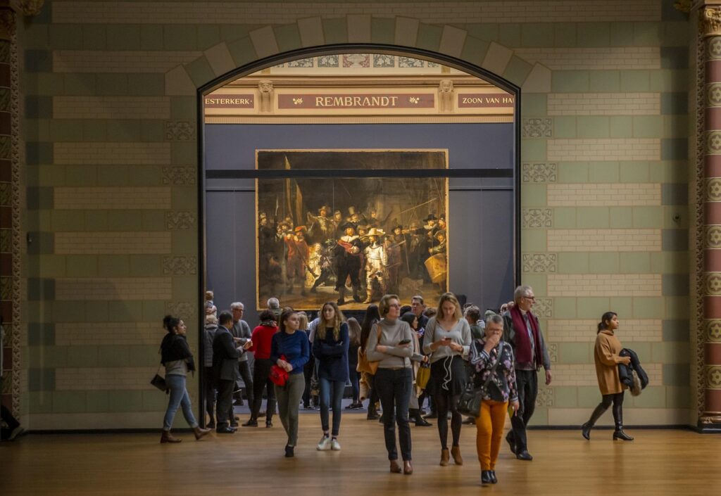 Date ideeën in Amsterdam 4 Een groep mensen, misschien op zoek naar hun ideale partner in de kunst, loopt en staat voor Rembrandts "De Nachtwacht", tentoongesteld in een grote, goed verlichte galerie met gedessineerde muren en een gewelfde deuropening.