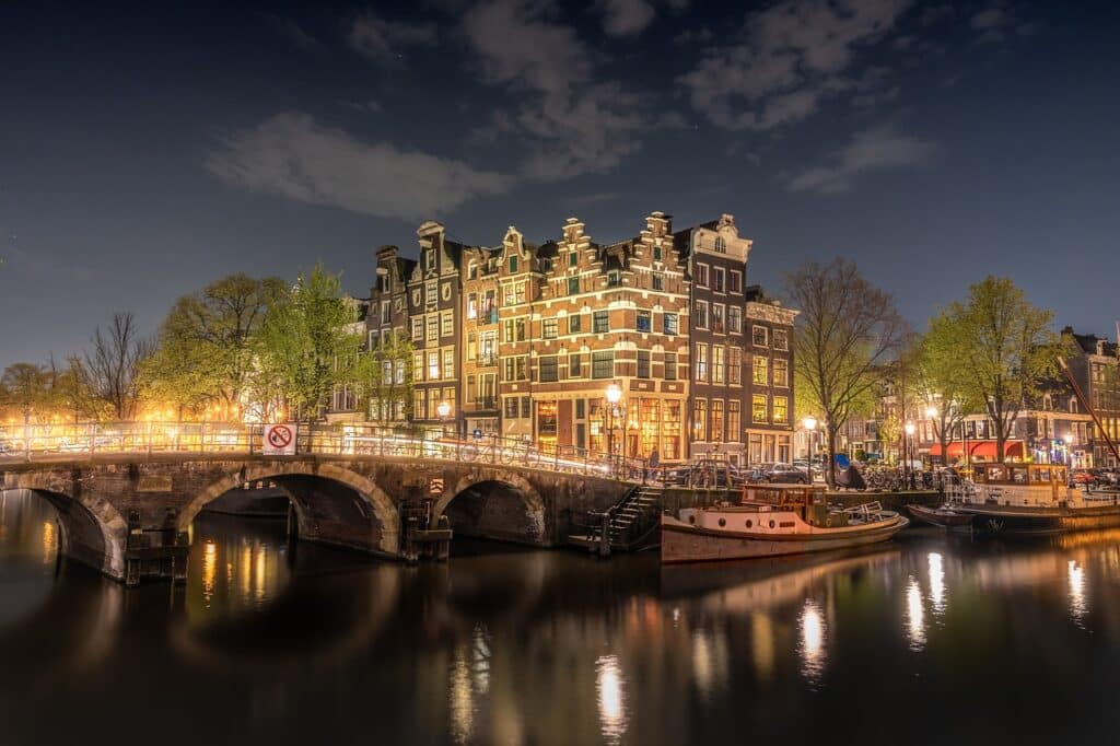 Date ideeën in Amsterdam 1 Nachtelijk uitzicht op de Amsterdamse gracht met een stenen brug, verlichte traditionele gebouwen met puntgevels en boten aangemeerd langs het water - een ideale partner voor een romantische wandeling onder bomen verlicht door straatverlichting onder een gedeeltelijk bewolkte hemel.