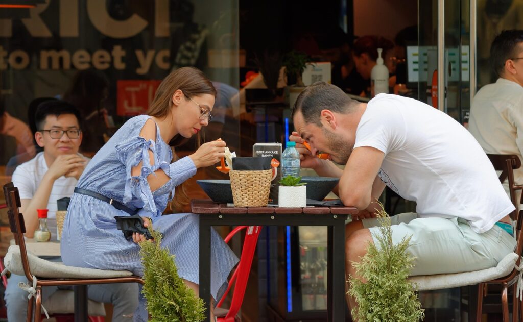 Date ideeën in Amsterdam 8 Een vrouw in een lichtblauwe jurk en haar ideale partner in een wit overhemd zitten aan een cafétafel in de buitenlucht, allebei geconcentreerd te eten uit een schaal, terwijl andere mensen op de achtergrond ontspannen.