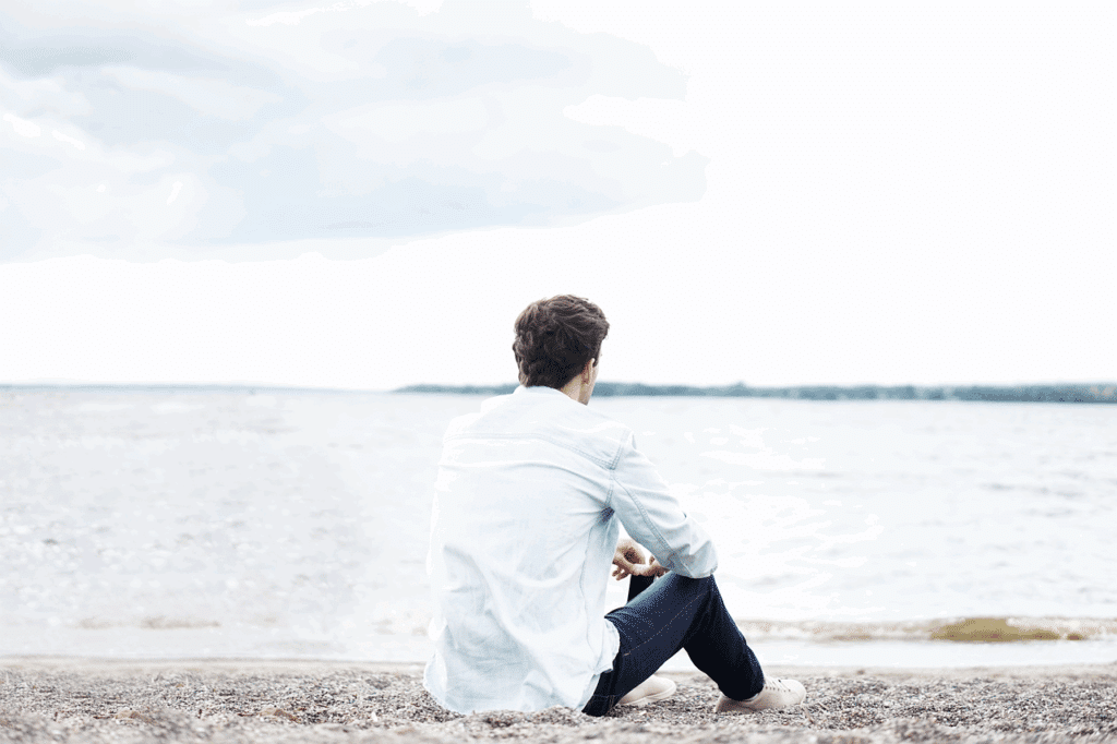 Een persoon met kort bruin haar, een licht overhemd en een donkere spijkerbroek, zit op een kiezelstrand met uitzicht op een kalm waterlichaam onder een bewolkte hemel, in stilte na te denken over de kwaliteiten van zijn ideale partner.