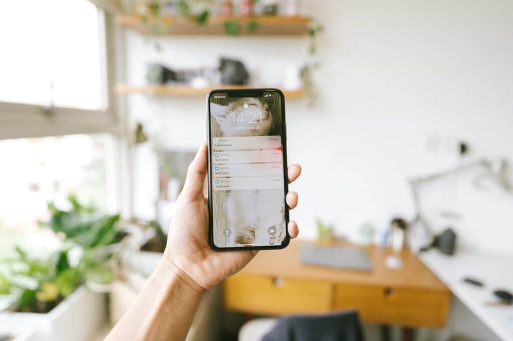 Een hand houdt een smartphone vast die meldingen weergeeft voor een thuiskantoor met een houten bureau, stoel, planten en planken - een ideale partner voor productiviteit in een lichte kamer vol natuurlijk licht.