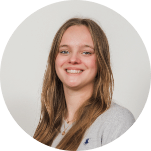 Een jonge vrouw met lang lichtbruin haar lacht naar de camera. Ze draagt een lichtgrijs topje en een gouden ketting tegen een effen witte achtergrond.