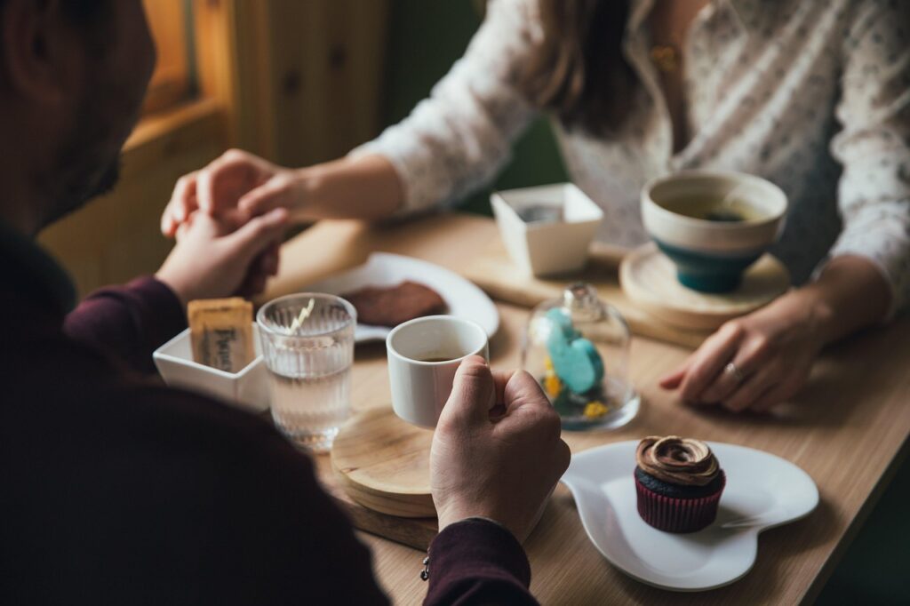 Een stel zit aan een houten tafel, houdt elkaars handen vast en deelt koffie, thee, een cupcake en gebak. De scène voelt warm en intiem aan en biedt inspiratie en datingtips voor wie op zoek is naar gezellige daten-momenten in natuurlijk licht.