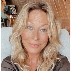 Een vrouw met golvend blond haar en blauwe ogen glimlacht zachtjes naar de camera terwijl ze binnen zit tegen een lichtgekleurde achtergrond, met houten muren zichtbaar achter haar.