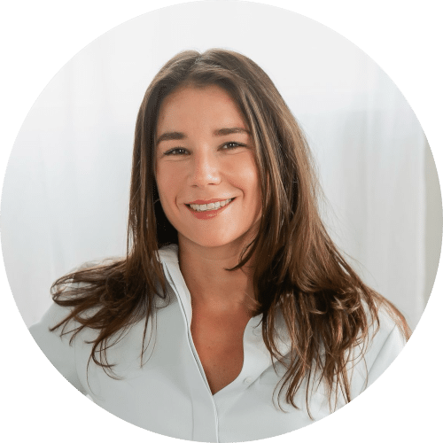 Een vrouw met lang bruin haar en een witte blouse lacht naar de camera terwijl ze voor een lichte achtergrond staat.