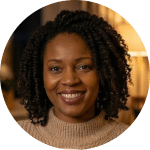 Een vrouw met krullend haar glimlacht warm en draagt een beige trui. In de zacht verlichte, gezellige binnensetting bladert ze door een aanbod en overweegt ze inschrijvingen voor komende evenementen.