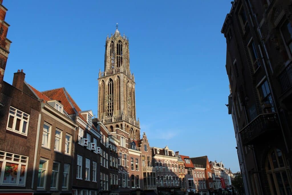 Een hoge kerktoren in gotische stijl steekt boven de rijen traditionele Nederlandse gebouwen uit onder een strakblauwe hemel in Utrecht - een romantische plek die perfect is voor een date-idee Utrecht.