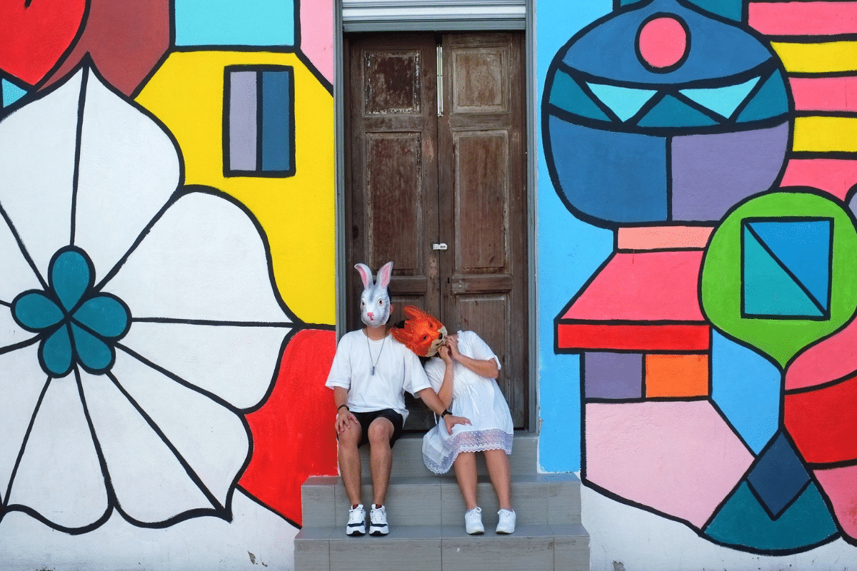 Tegenpolen – stel graffiti Twee mensen, gekleed in het wit en met tegenpolenmaskers - een konijn en een vos - zitten op een trapje voor een houten deur, omlijst door levendige geometrische straatkunst op de muur achter hen.