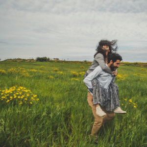 Een man geeft een vrouw een ritje op zijn rug door een veld met groen gras en gele bloemen. Beiden lachen en hun lach laat zien hoe zelfs tegenpolen samen vreugde kunnen vinden terwijl het haar van de vrouw in de wind waait onder een bewolkte hemel.