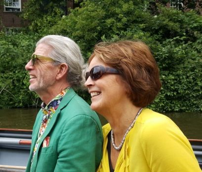 Succesverhalen - Start 11 Twee oudere volwassenen, een man en een vrouw, zitten buiten aan een rivier, allebei glimlachend en met een zonnebril op. De man heeft grijs haar, een baard en draagt een groene blazer met een kleurrijk overhemd. De vrouw heeft kort bruin haar en draagt een geel topje met een ketting. Ze hebben elkaar ontmoet via een deskundige matchmaking-service.