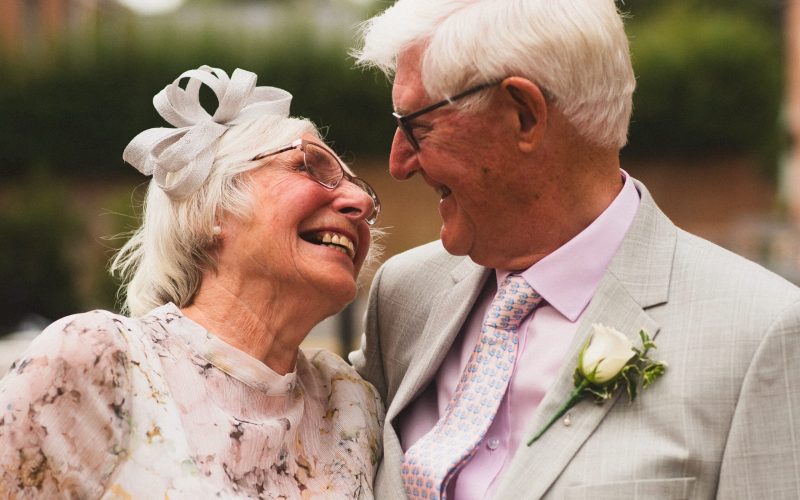 Een ouder echtpaar, formeel gekleed, met de vrouw in een gebloemde jurk en fascinator, en de man in een lichtgrijs pak met een boutonnière, lachen vrolijk en kijken elkaar aan. De achtergrond van groen en een bakstenen gebouw roept het succes op van matchmaking via een datingbureau.