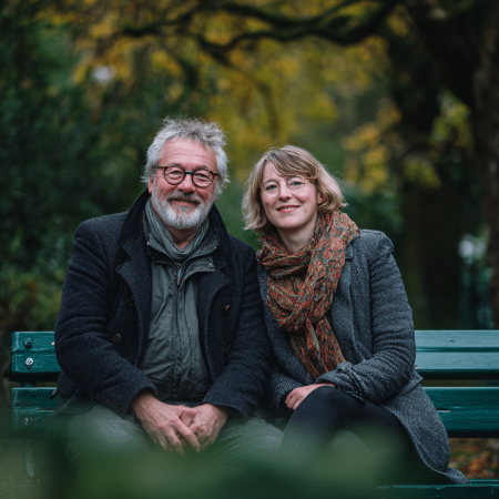 Een oudere man en een jongere vrouw in een leeftijdsverschil zitten dicht tegen elkaar aan op een groen bankje in het park, lachend naar de camera. Beiden dragen jassen en sjaals, met vage herfstbomen en groen op de achtergrond.