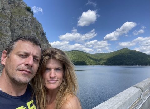 Succesverhalen - Start 1 Een man en een vrouw staan dicht bij elkaar en maken een selfie met een schilderachtige achtergrond. Achter hen bevinden zich een grote rotsformatie, een watermassa en glooiende groene heuvels onder een blauwe lucht bezaaid met wolken - momenten prachtig vastgelegd terwijl ze hun duurzame relatie vieren.